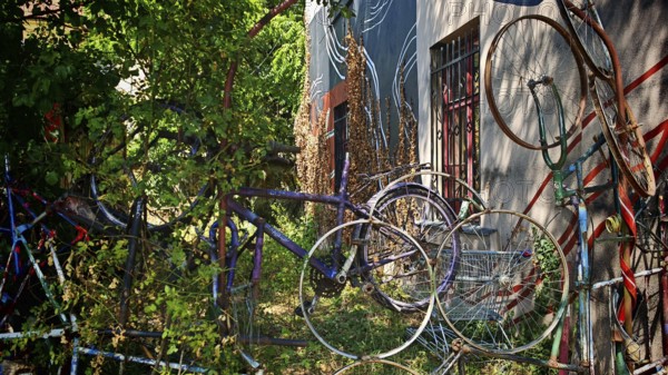 Colourful bicycles stacked on top of each other next to an art-painted wall surrounded by plants,