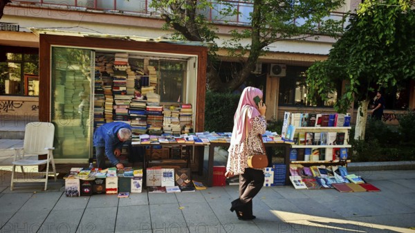 Book stand on a street surrounded by people and green trees, idyllic street scene with woman