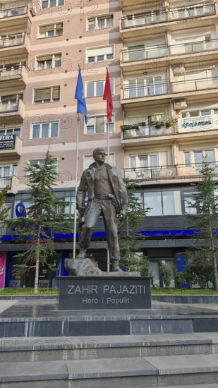 Statue of Zahir Pajaziti in front of a modern building with two flags in the background, Pristina,