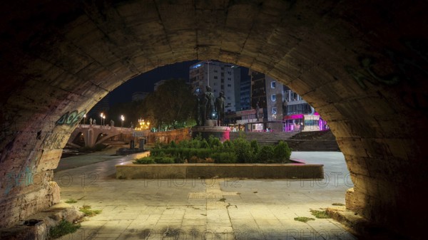 View through a stone archway of an illuminated square with monuments at night, Skopje, North