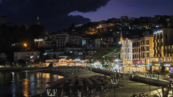 A nighttime beach view with illuminated buildings and dark skies in a lively coastal town, Ulcinj