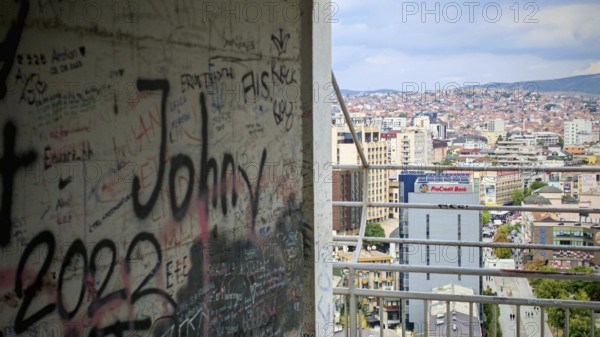A graffiti-covered building offers a view of an urban landscape under a cloudy sky, Pristina,