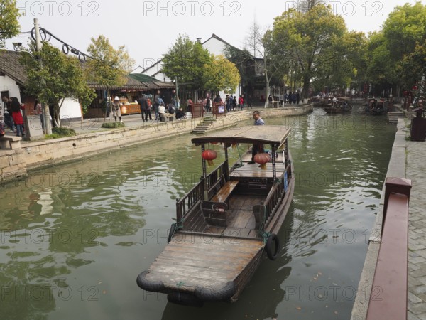 A wooden boat floats on a canal next to traditional buildings and market visitors, Zhujiajiao