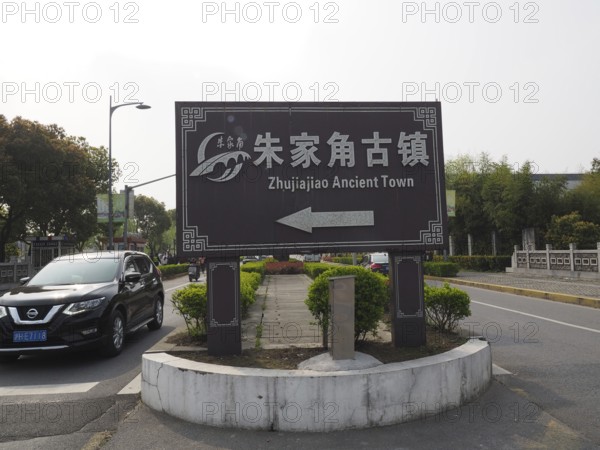 A road sign points to the ancient city of Zhujiajiao surrounded by trees and a road with cars,
