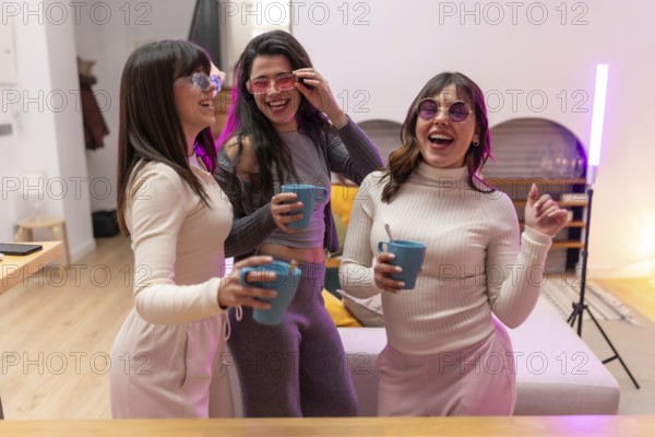 Three cheerful young female friends wearing sunglasses, laughing and holding hot drinks in mugs,