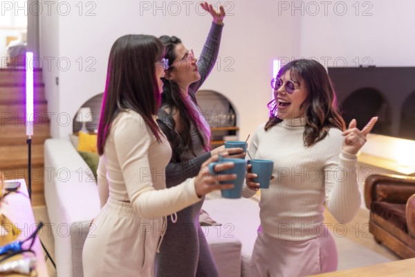 Three young women laughing and celebrating with drinks in a cozy apartment living room, purple pink
