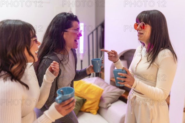 Three young women laughing and chatting over hot drinks in a cozy apartment, enjoying a relaxed