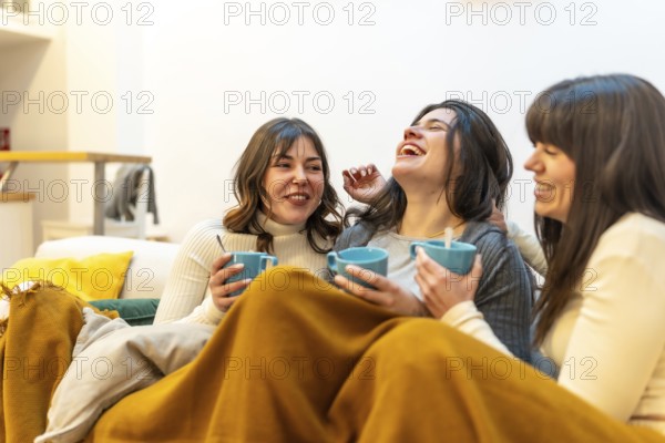Happy women friends enjoying a cozy moment together at home, laughing and having hot drinks while