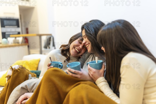 Three young women relaxing on a sofa at home, wrapped in a blanket, laughing and sharing warm