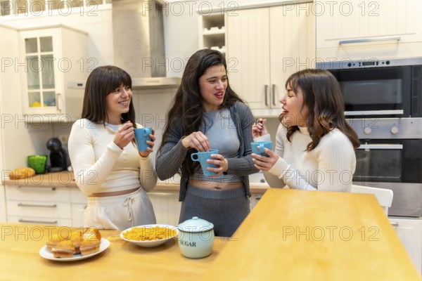Three young women laughing and chatting around a kitchen island, sharing muffins and hot drinks in