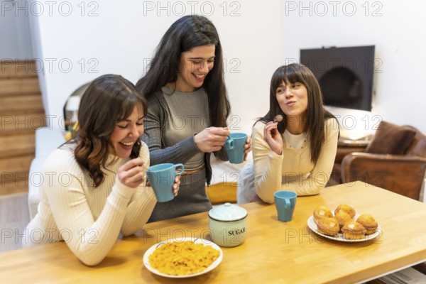 Three young women smiling and laughing, holding cups and spoons, gathering around a wooden kitchen
