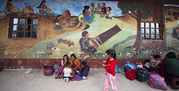 Mayan families in traditional clothes squat in front of a mural at the market in Chichicastenango,