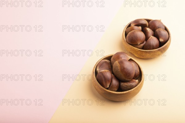Wooden bowl with raw edible ?hestnuts on pink and orange paper pastel background, side view, copy