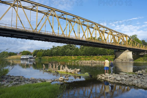 Yellow metal bridge across river with two anglers on the shore and blue sky, bridge over Morava