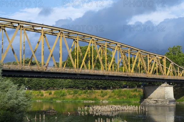 Yellow metal bridge over a river with green vegetation on the banks and cloudy sky, bridge over the