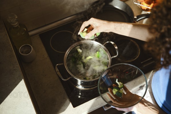 Fresh broccoli is placed into a pot of boiling water on the stove. A woman stands over the pot,