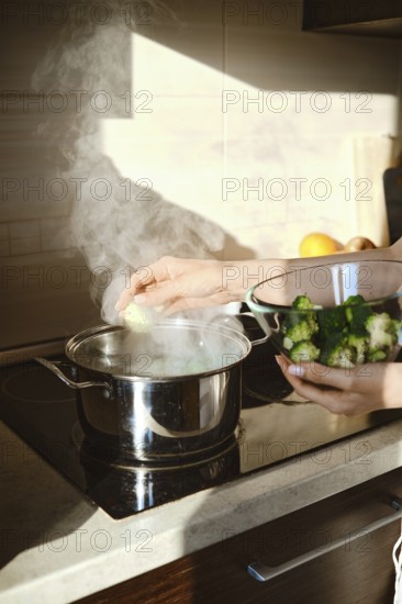 A person is cooking broccoli in a pot on the stove. Steam rises from the pot while another bowl