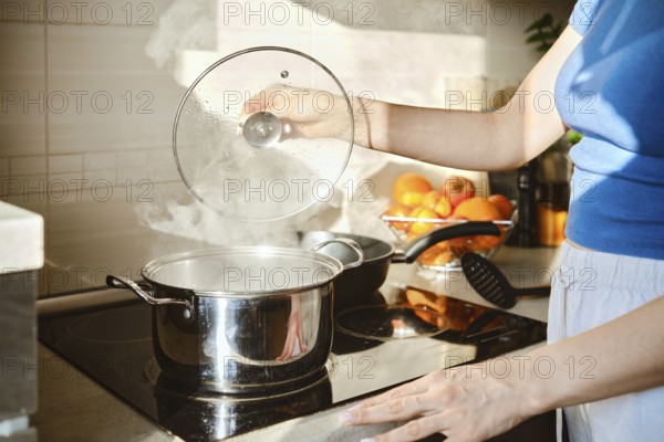 A woman lifts the lid of a saucepan in which water is boiling. Hot steam rises from the pan and is