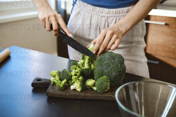 Close-up view of cutting fresh broccoli on a wooden board in a home kitchen. The person uses a