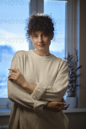 A young woman stands near a window in a dim room. She is wearing a light sweater and looks at the