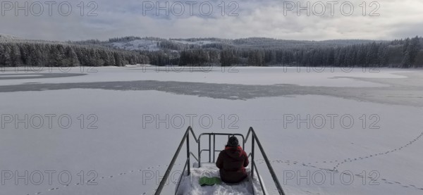 Person sitting on snowy footbridge with a view of the partially frozen lake, Rennsteig, Thuringian