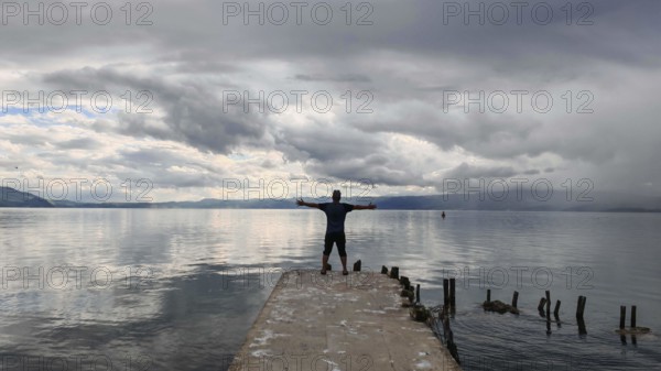 Person standing with outstretched arms on a pier surrounded by water and clouds, Lake Ohrid, North