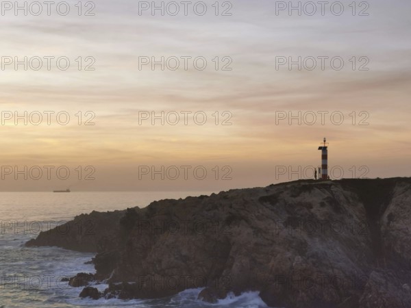 Landscape with lighthouse on rocks, illuminated by sunset, with calm sea and clouds in the