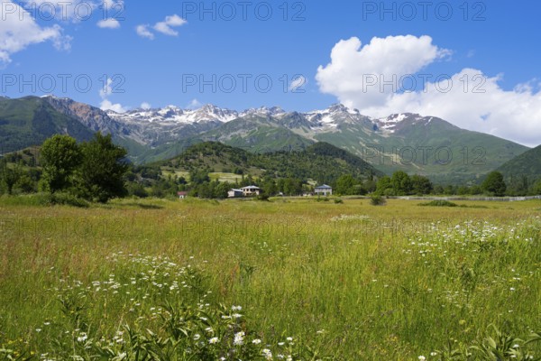 Wide meadow landscape with views of mountains and cloudy blue sky, surrounded by green hills,