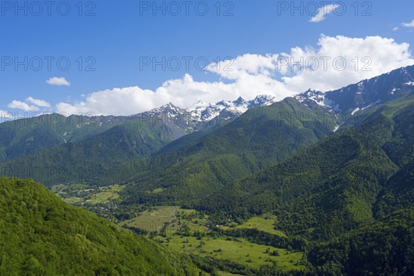 Green valley with densely wooded slopes and distant mountains under blue sky with clouds, landscape