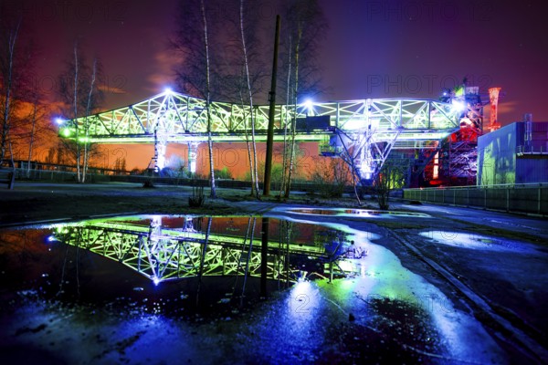 Illuminated bridge at night with blue and green lighting reflecting in a pool of water,