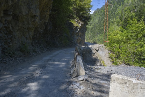 A narrow mountain road leads between rocky cliffs and green forest. Sun and shade areas alternate,