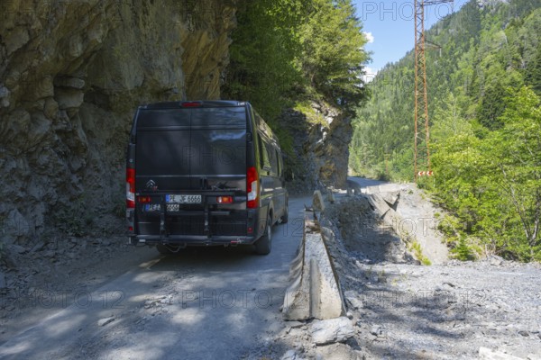 A black van drives on a narrow mountain road surrounded by rocks and thick vegetation, camper