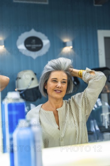 Mature woman looking at her reflection, touching her fresh gray hairstyle, and smiling in a modern