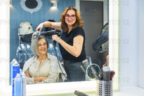 Female hairdresser smiling at camera while blow drying a mature customer's hair in a modern salon,