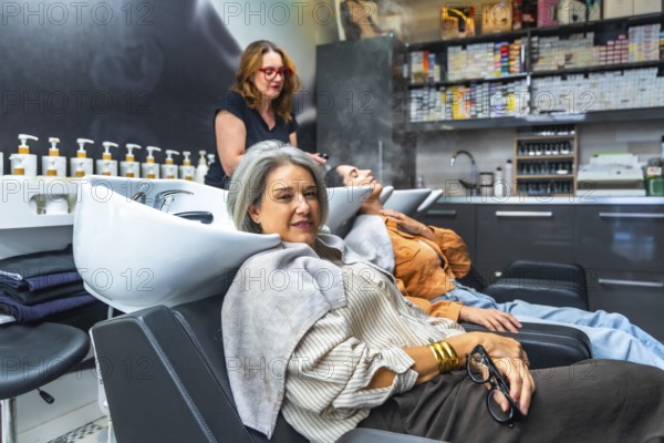 Two women reclining at salon basins while a stylist performs professional shampoo and scalp care,