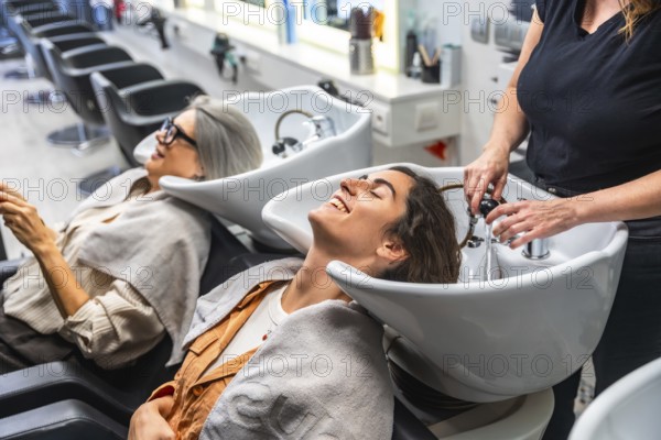 Female hairdresser washing a young woman's hair in a salon hair wash basin, while another client