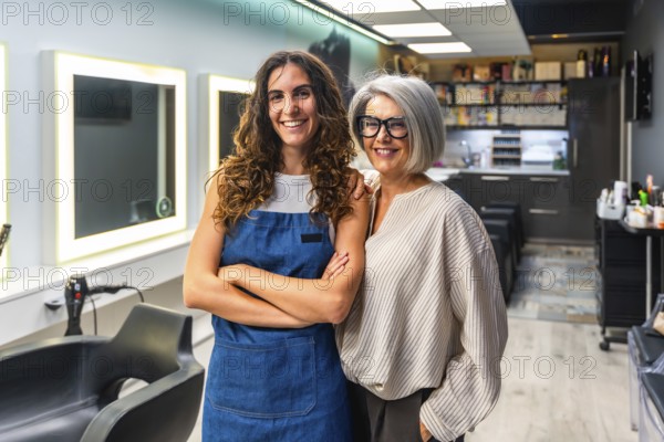 Two cheerful women hairstylists, one younger in an apron and one older with gray hair, standing