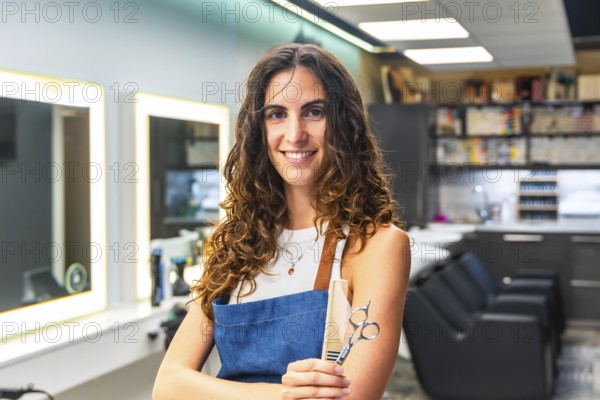 Professional woman with curly hair wearing an apron and holding hairdressing scissors and a comb,