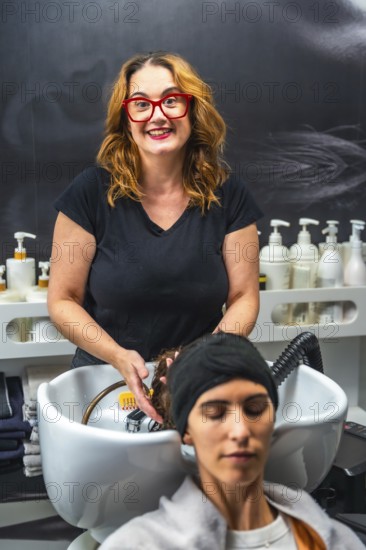 Hairdresser smiling while washing client's long red hair over salon sink, providing professional
