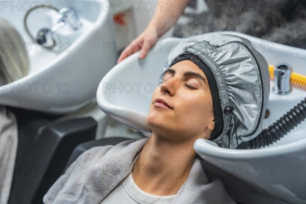 Woman lying at a hair washing sink, receiving a moisturizing hair steaming treatment from a