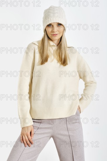 A model stands confidently in a studio while wearing a light sweater and a knitted hat. The setting