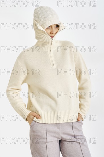 A woman stands in a neutral background wearing a warm knitted sweater and a hat. She looks relaxed