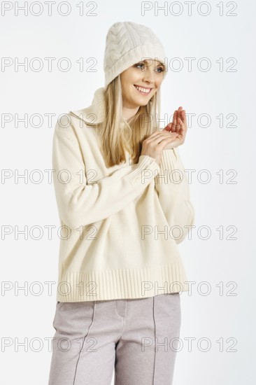 A woman stands with a smile, wearing a sweater and a hat. She clasps her hands together in front of