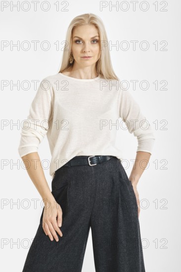 A woman stands with a neutral expression in a studio. She wears a light top and gray wide pants.
