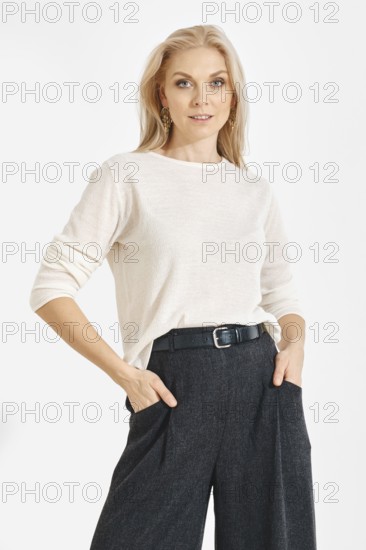 A woman poses with hands in pockets wearing a light shirt and dark pants. She has long blonde hair