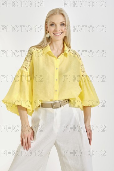 Cheerful woman in a vibrant yellow blouse with lace details and white trousers posing in bright