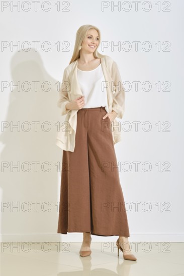 A smiling woman stands gracefully in a bright, minimalist studio, dressed in a light top and