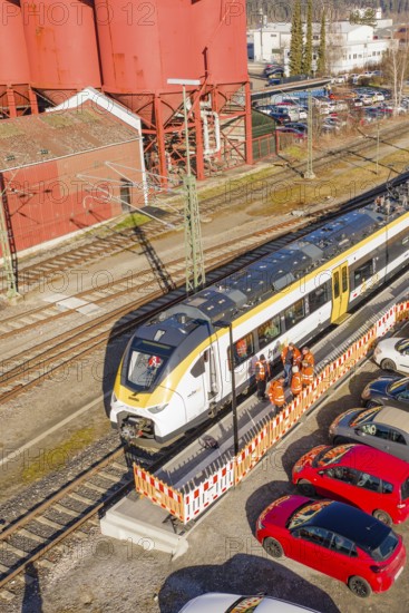 Close-up of a train at a station surrounded by industrial buildings, cars and several workers, test