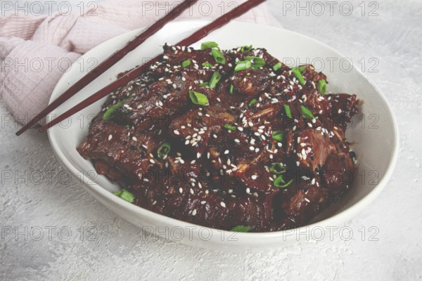 Crispy beef, with green onions and sesame seeds, in a bowl with chopsticks, on a textured surface,
