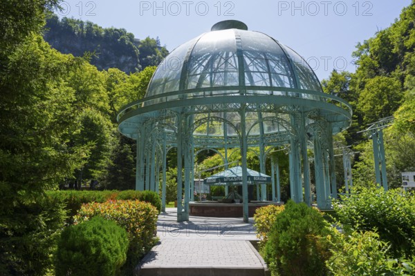 An elegant glass and metal pavilion in a green, sunny landscape surrounded by trees, pavilion with